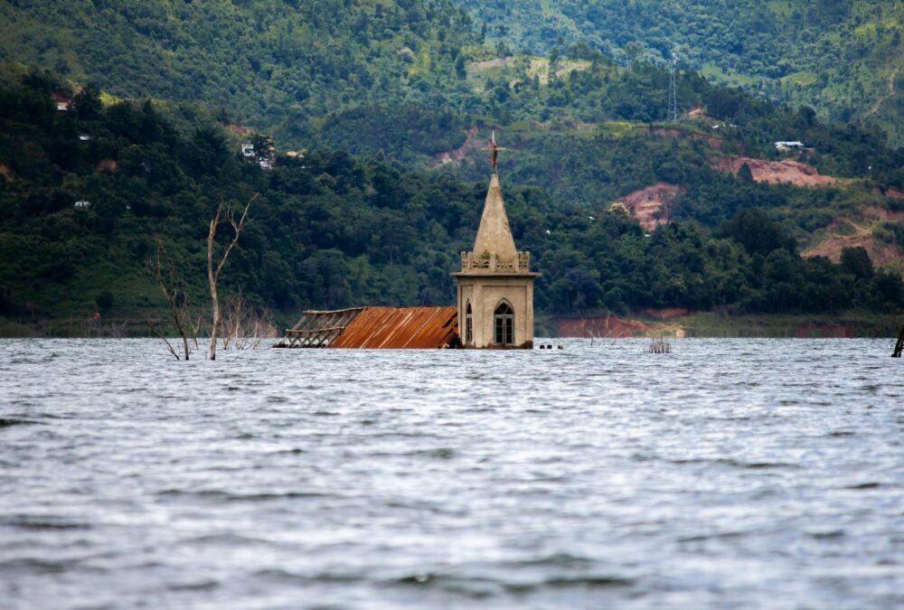 Photo of the spire and roof of a building above flooded waters with mountains in the background.