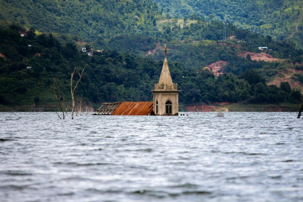 Photo of the spire and roof of a building above flooded waters with mountains in the background.